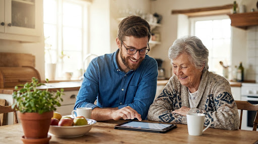 A friendly technician sitting at a sunlit kitchen table with an elderly woman, gently guiding her through using a tablet.